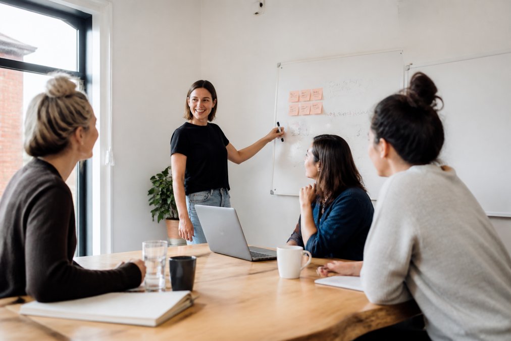A small group of business founders seated around a table in a bright meeting room, one person facilitating at a whiteboard with notes and a laptop open