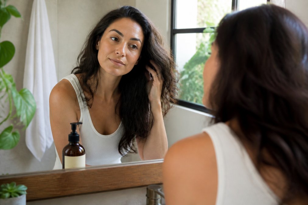 Woman examining her hair in a bathroom mirror while transitioning to an organic haircare routine.