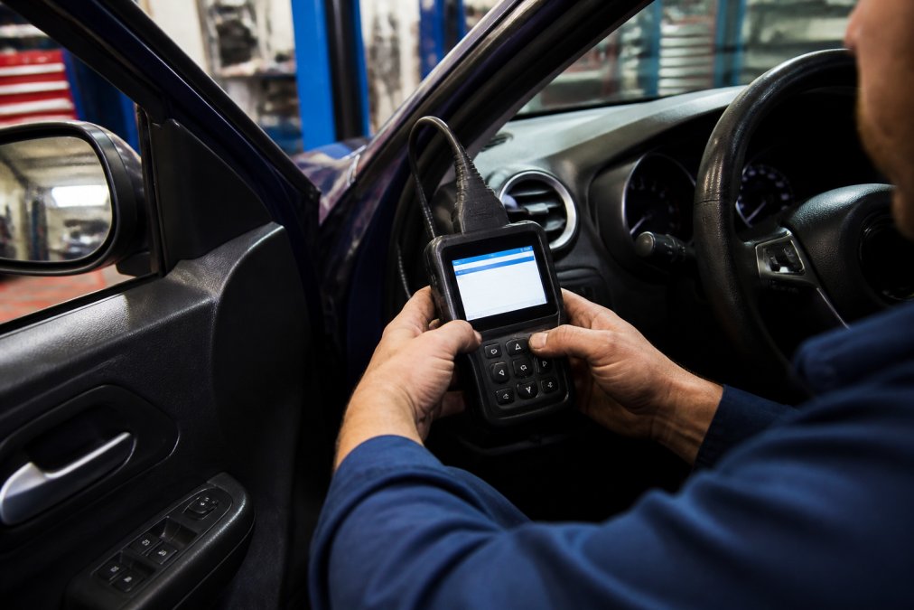 Close-up of a mechanic's hands holding a diagnostic scanner connected to a vehicle's onboard computer system, with the scanner screen displaying fault codes in a modern repair bay