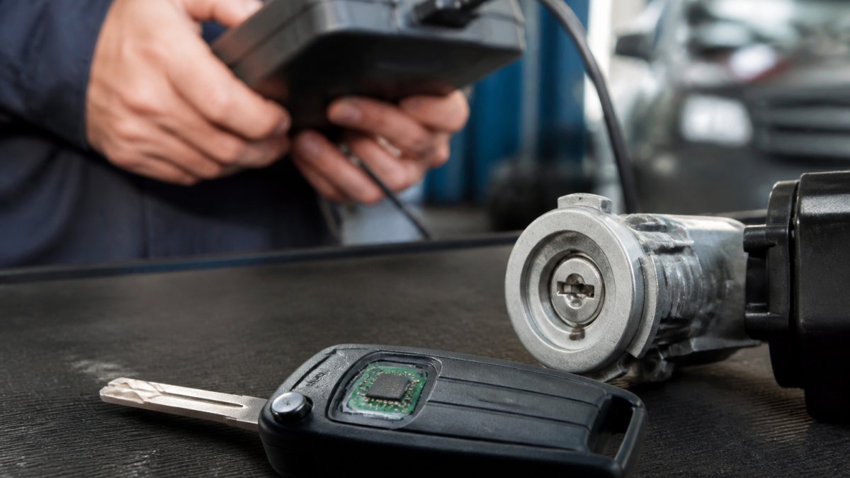 Close-up of a car key with a visible transponder chip resting beside an ignition cylinder, a locksmith's hands holding a diagnostic scan tool in a dimly lit garage