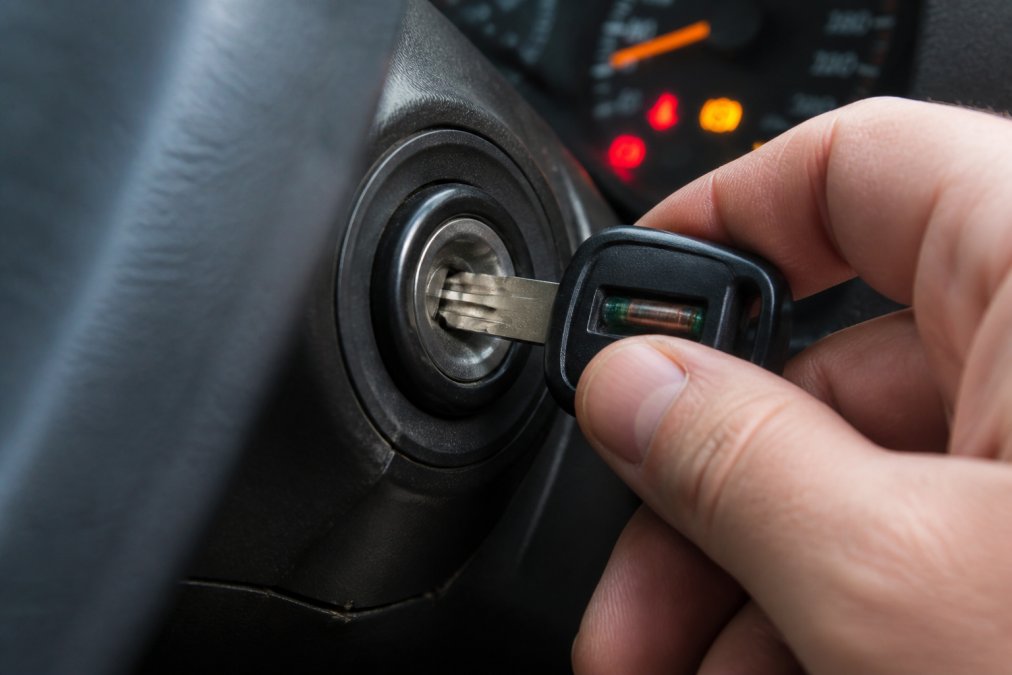 Close-up of a hand inserting a transponder key into a car ignition cylinder, showing the key blade and plastic head with visible microchip area, dashboard lights glowing softly in the background