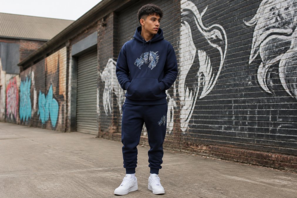 Young man in his early twenties wearing an oversized graphic hoodie in navy with matching navy joggers and clean white trainers, standing in an urban setting with brick walls and street art visible in the background, demonstrating a complete budget streetwear outfit with intentional styling