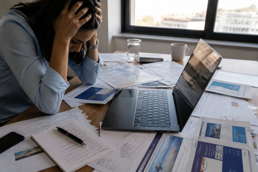 Stressed traveler at a laptop surrounded by open browser tabs showing multiple airline and hotel booking sites, printed itineraries, and handwritten notes scattered across desk, natural afternoon light from office window