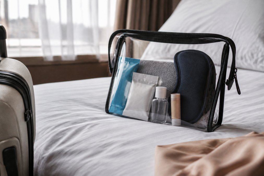 A flat-lay photograph of a carry-on wellness kit on a hotel bed, showing a transparent pouch of travel-sized items including a hydration stick pack, a small patch pouch, hand sanitizer, lip balm, and a sleep mask, with soft natural light from a nearby window