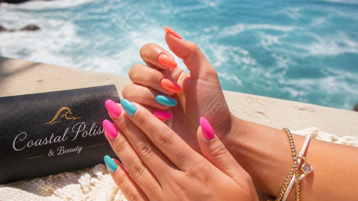 Close-up of hands with vibrant summer nail polish shades, turquoise, coral, and neon pink, against a coastal backdrop with ocean water visible in the background