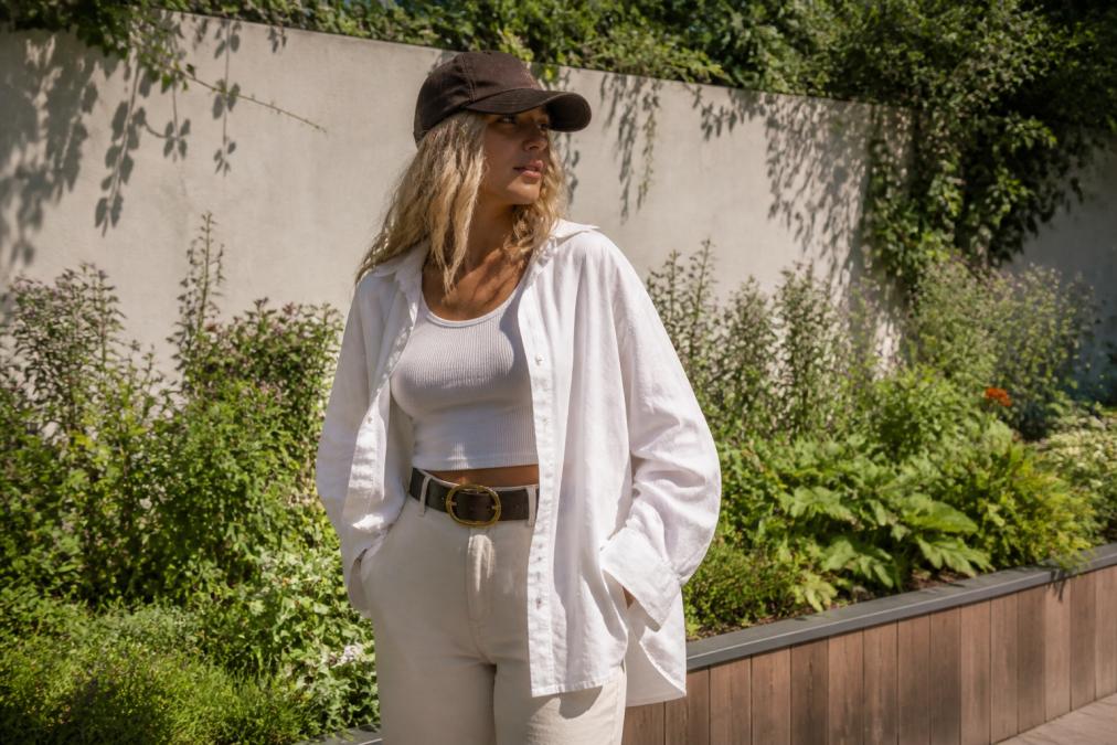 Person trägt ein oversized Leinenhemd über einem Tanktop, dazu einen auffälligen Gürtel und eine Cap, in natürlichem Sonnenlicht vor einem sommerlichen Gartenhintergrund