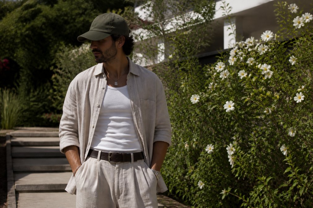 Person in trendiger Streetwear mit einem Oversize-Leinenhemd und einer Kappe in einem sonnigen Sommergarten.
