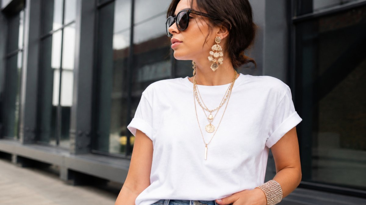 Woman wearing a simple white t-shirt and jeans, styled with layered gold necklaces, oversized dangling earrings, and a wide rhinestone cuff bracelet, photographed against a neutral background to showcase how accessories transform the outfit