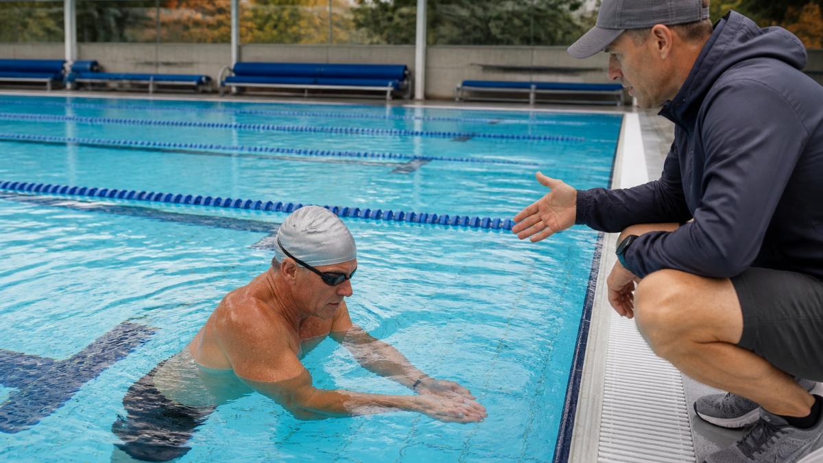 A masters athlete in their 50s performing a sculling drill in a lap pool, with a coach on the pool deck observing and offering feedback