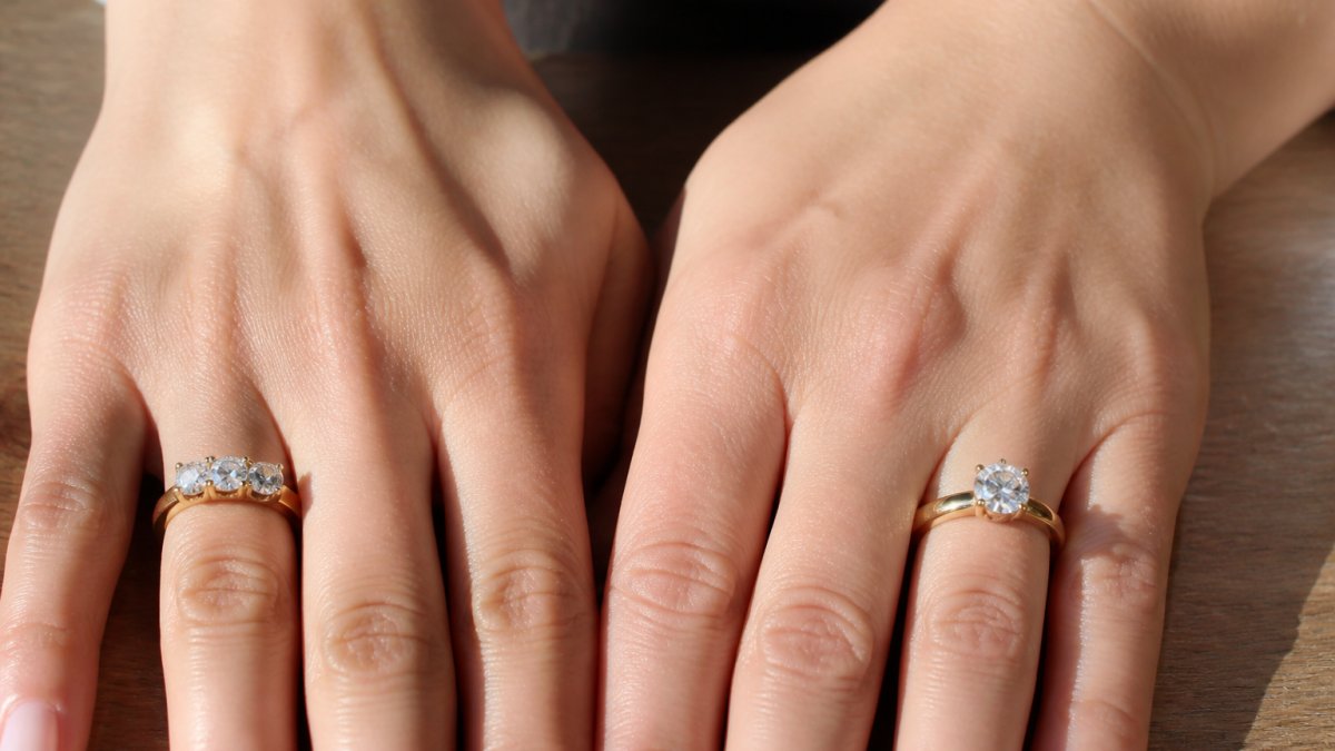 Hands wearing an 18k yellow gold trilogy ring and a single-stone solitaire ring on a wooden surface