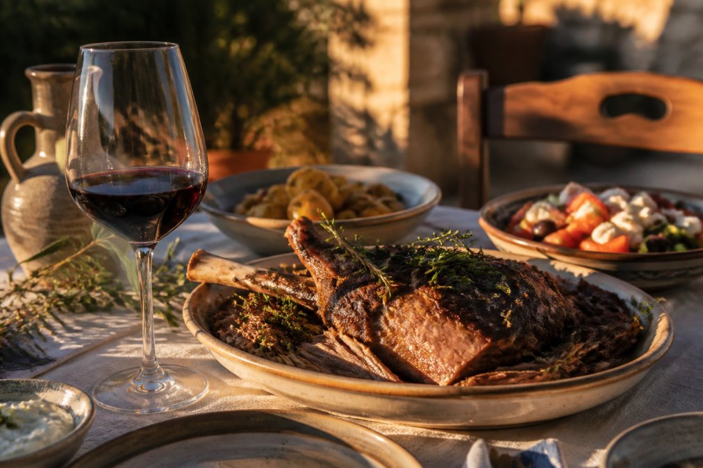 Ein gedeckter Tisch im mediterranen Stil mit gegrilltem Lammfleisch auf einer Platte, einem Glas dunkelrotem Wein daneben und frischen Rosmarin- und Thymianzweigen, warmes Abendlicht von der Seite