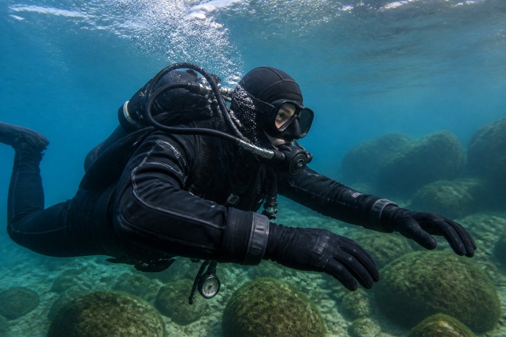 Tauchprofessional in Trockentauchanzug unter Wasser, klare Luftschicht sichtbar um Körper und Arme, helles Bodensee-Wasser mit Felsen im Hintergrund