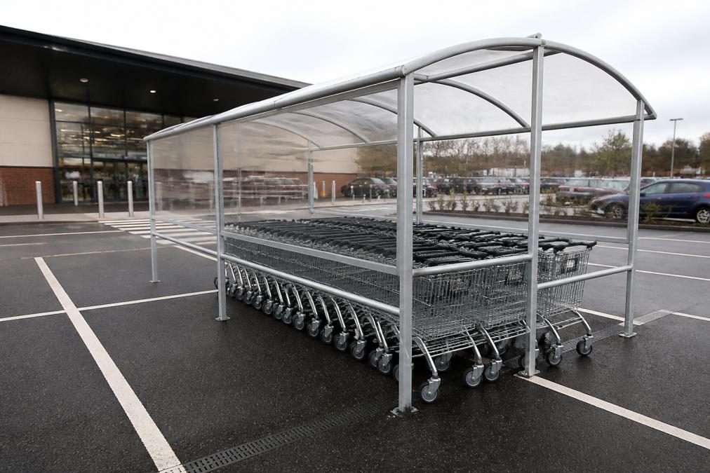 A row of supermarket shopping trolleys parked neatly inside a galvanised steel open-fronted trolley shelter in a car park, with a store entrance visible in the background on an overcast day
