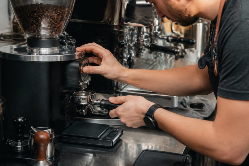 Close-up of a barista's hands adjusting the grind size on a burr grinder, with a portafilter and digital scale nearby on a stainless steel countertop