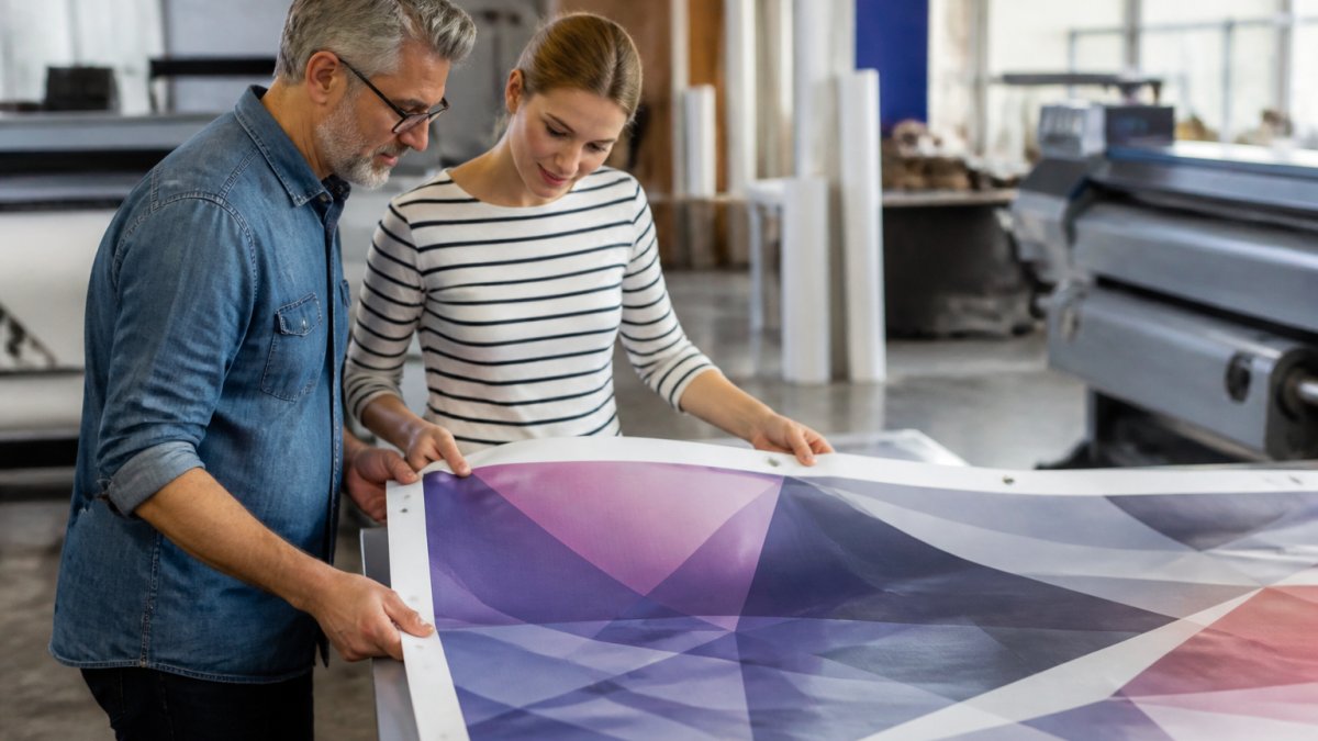 A print production specialist and a client reviewing a hard copy proof of a branded banner next to a monitor showing the same artwork, in a bright print shop