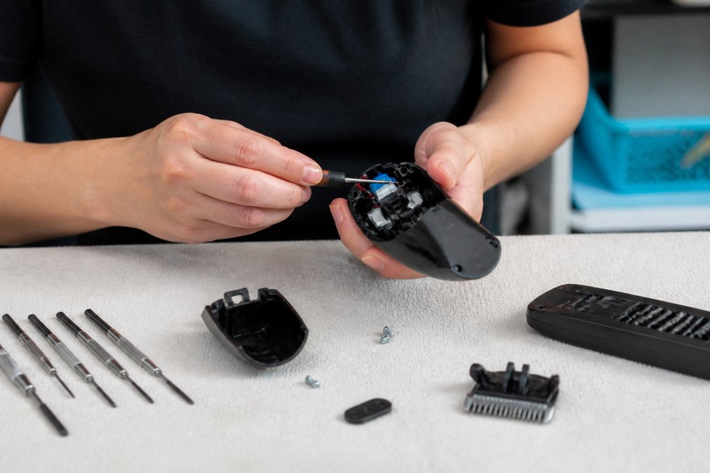 Close-up of a person's hands using a small screwdriver to open a dog clipper housing, with a clean workspace and precision tools laid out on a light-colored table