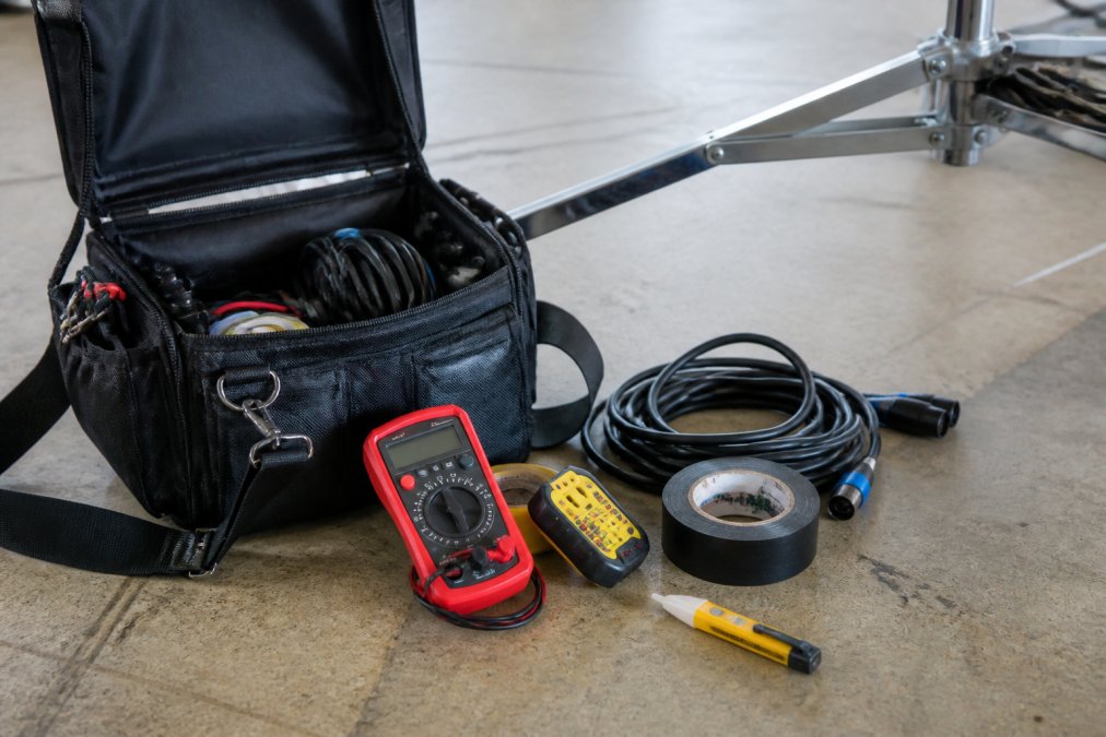A lighting technician's open tool bag on a film set floor, showing a multimeter, spare DMX cables, gaffer tape, and a continuity tester laid out beside a light stand