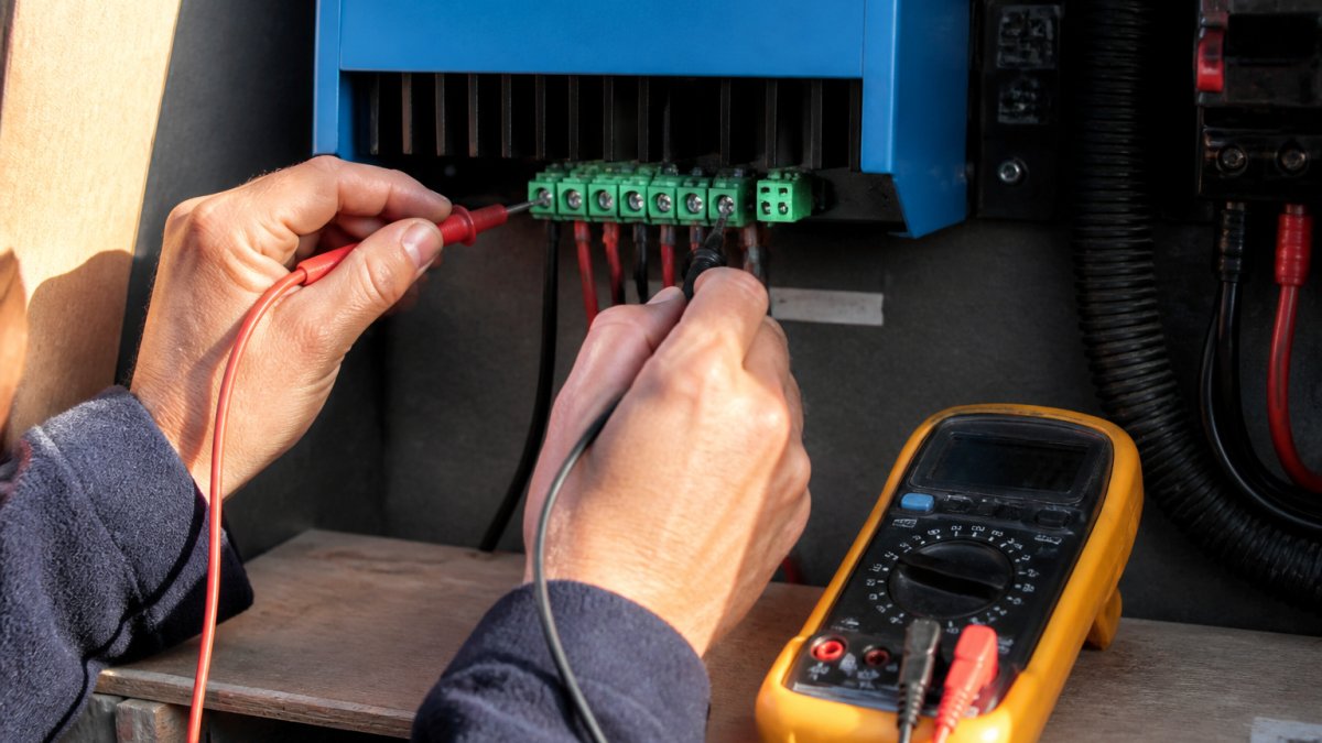 Close-up of a technician's hands using a digital multimeter to test voltage connections on a solar charge controller terminal block in bright natural light