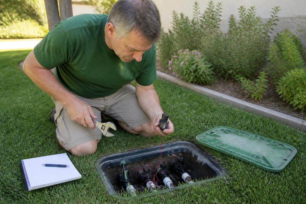 A homeowner kneeling beside an open irrigation valve box in a green lawn, holding a wrench and inspecting a solenoid with a notepad nearby