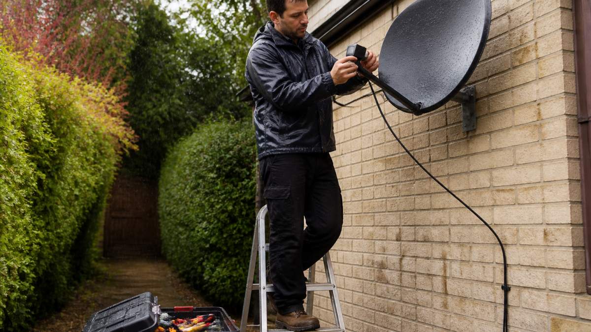 Troubleshooting Satellite Signal Loss in Heavy Rain A person on a stepladder in a garden, carefully inspecting the cable connections on the back of a satellite dish during light rain, with a toolbox nearby