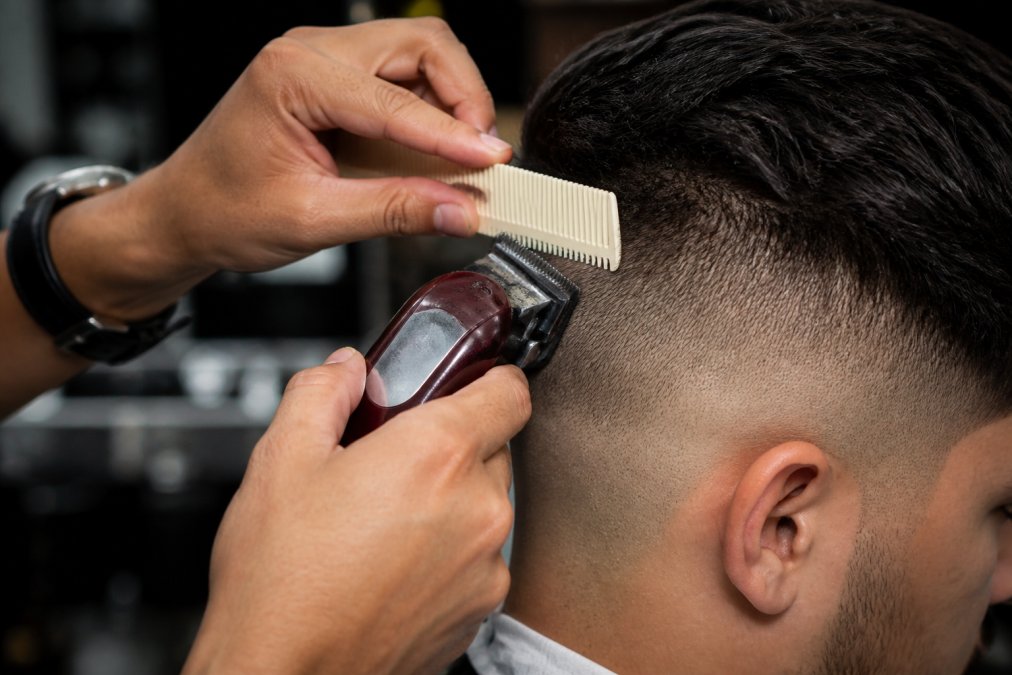 Close-up of a barber's hands using clippers with a fade comb to blend and smooth a harsh line on a client's head, showing precise clipper-over-comb technique and proper blade angle