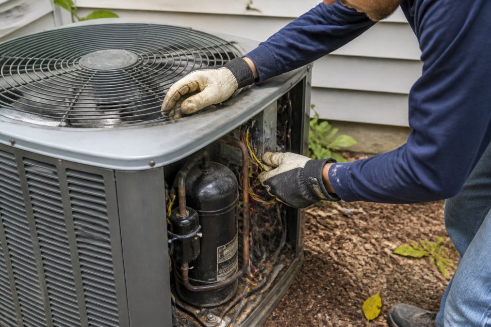 Close-up of a homeowner's hands wearing protective gloves inspecting an outdoor AC unit's condenser fan and compressor area, checking for debris and loose components in bright daylight