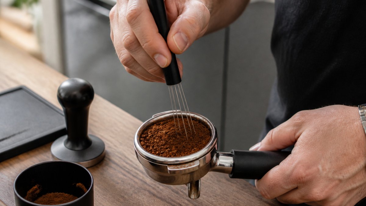 Hands using a needle distribution tool to level coffee grounds in a portafilter basket on a wooden counter.