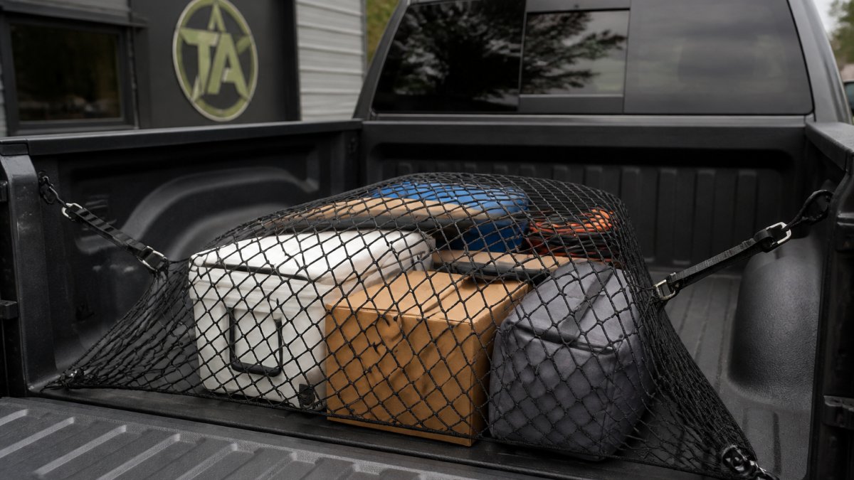 Close-up of a truck bed showing a cargo net stretched across with bed hooks visible on the sides and tie-down straps taut, securing mixed items including boxes and equipment