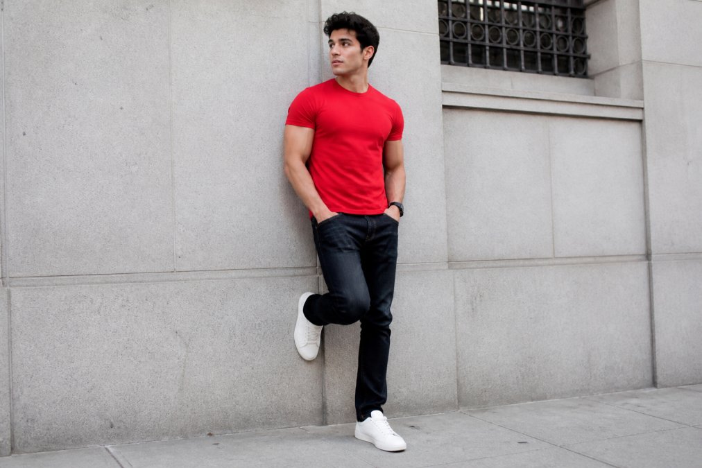 Young adult wearing a bright red True Religion red shirt with dark jeans and sneakers against a city wall