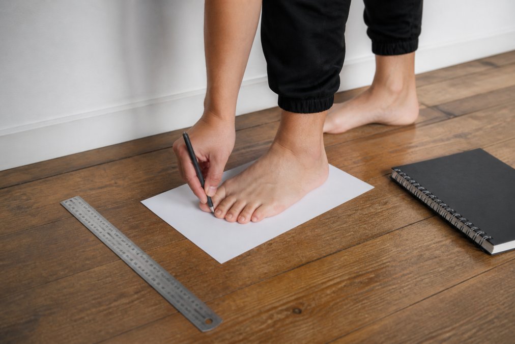 A person standing barefoot on a sheet of paper against a wall, marking the heel and longest toe with a pen, with a ruler and notebook nearby on a wooden floor