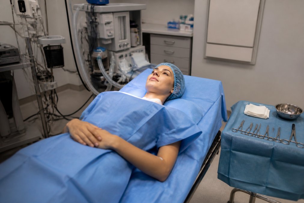 Patient in surgical attire lying on operating table in clean, well-lit surgical suite with monitoring equipment and surgical instruments visible, demonstrating a calm, professional medical environment
