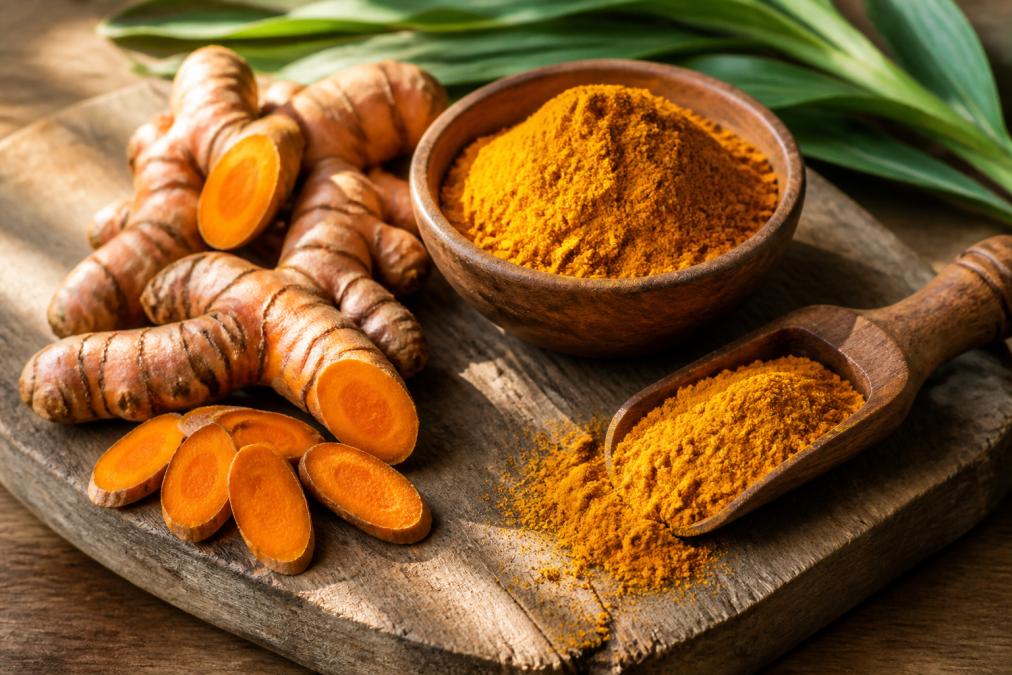 Close-up of fresh golden turmeric root and ground turmeric powder arranged on a weathered wooden cutting board with natural sunlight streaming across the surface, emphasizing the vibrant golden-orange color and texture
