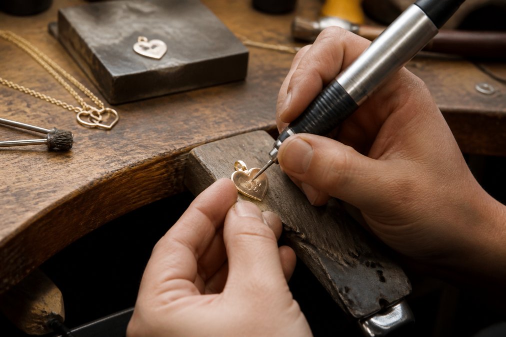 Close-up of hands carefully engraving a piece of jewelry on a workbench with crafting tools, precision instruments, and personalized designs visible under bright workshop lighting