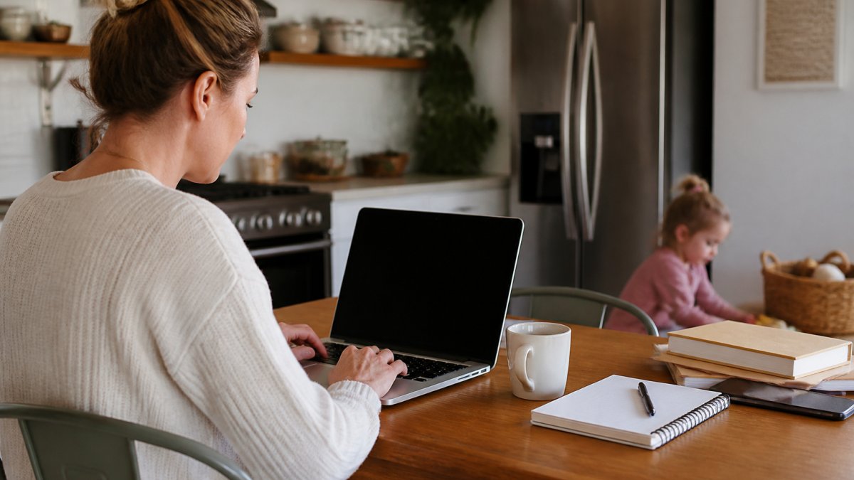 A parent working at a kitchen table with a laptop, notebook, and coffee, setting up an online business dashboard while a child plays nearby