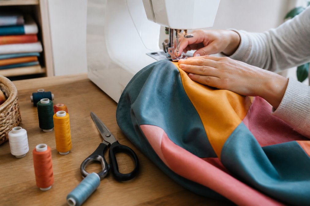 Mains cousant une housse de pouf colorée sur une machine à coudre, avec des bobines de fil et des ciseaux posés sur la table de travail