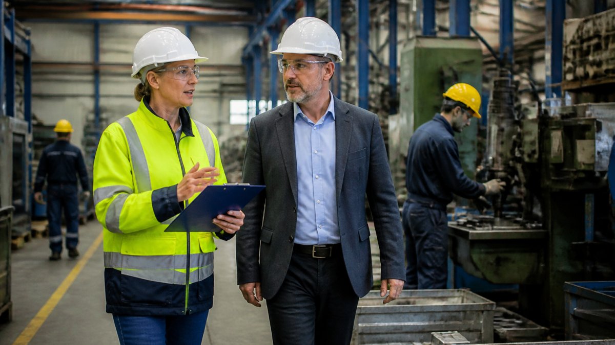Eine Fachkraft für Arbeitssicherheit mit Warnweste und Klemmbrett führt gemeinsam mit einem Betriebsleiter eine Sicherheitsbegehung in einer Produktionshalle durch, im Hintergrund laufen Maschinen und beschäftigte Mitarbeiter tragen Schutzausrüstung