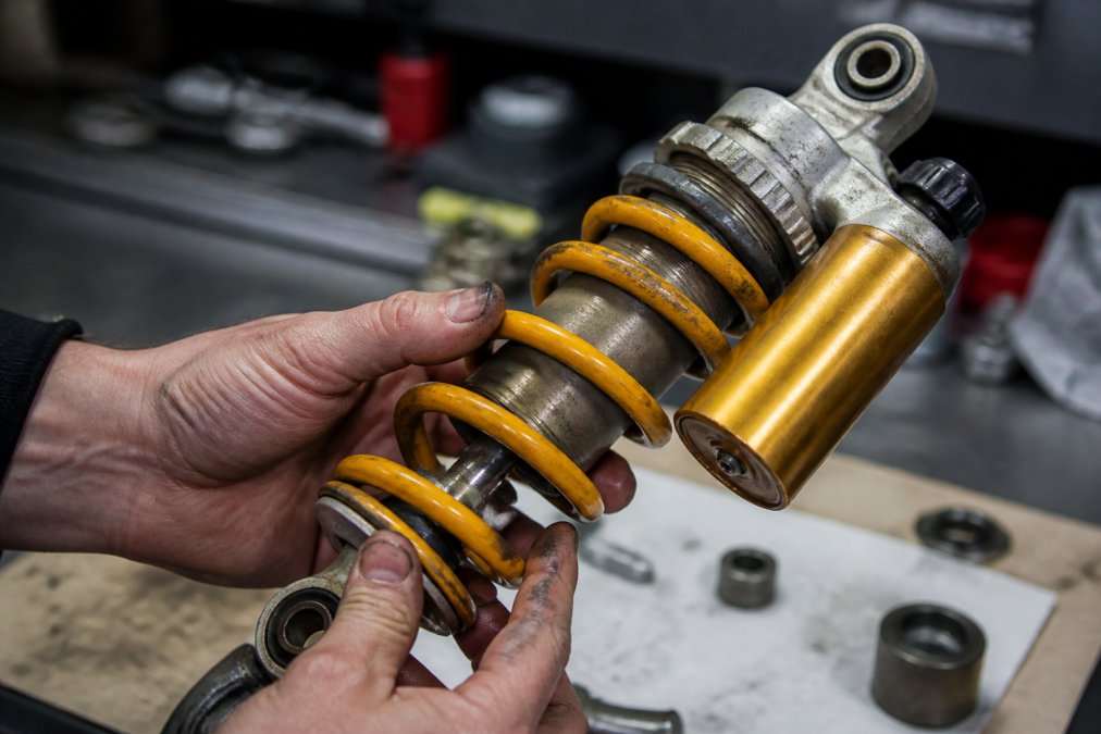 Close-up van handen die een Öhlins-schokdemper inspecteren op slijtage, met duidelijk zicht op de demper en beschadigingen onder werkplaatslicht