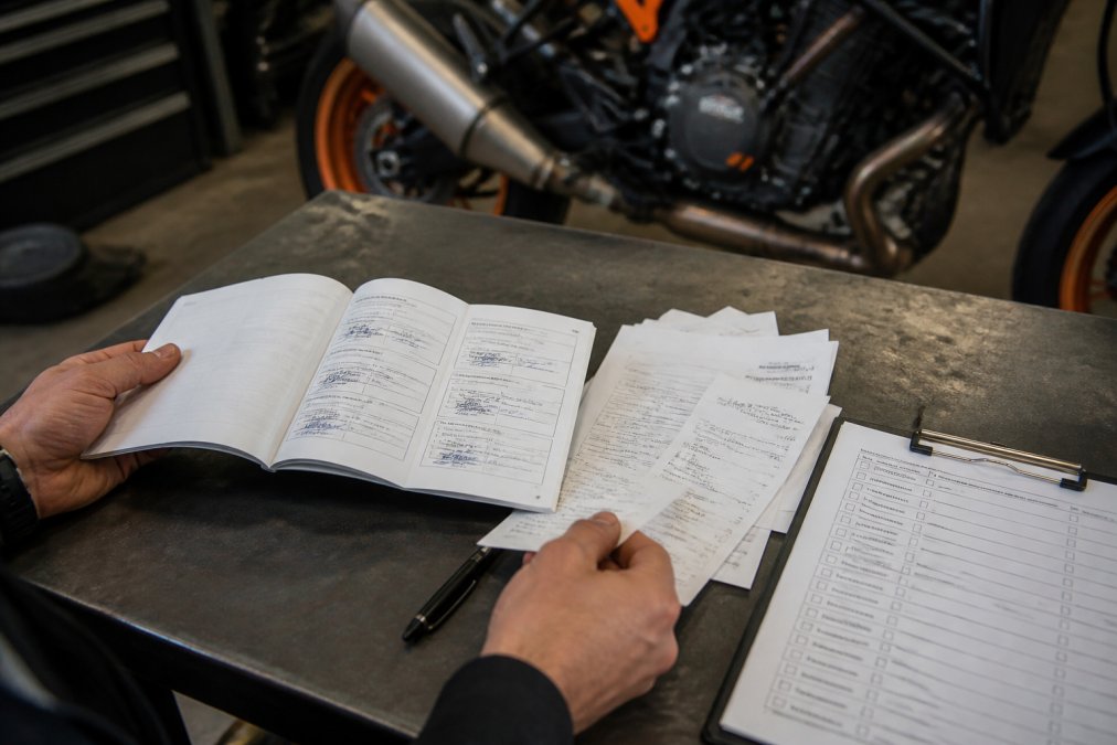 Close-up van handen die een motorfiets onderhoudsboekje en facturen doornemen op een werkbank, met een checklist naast zich