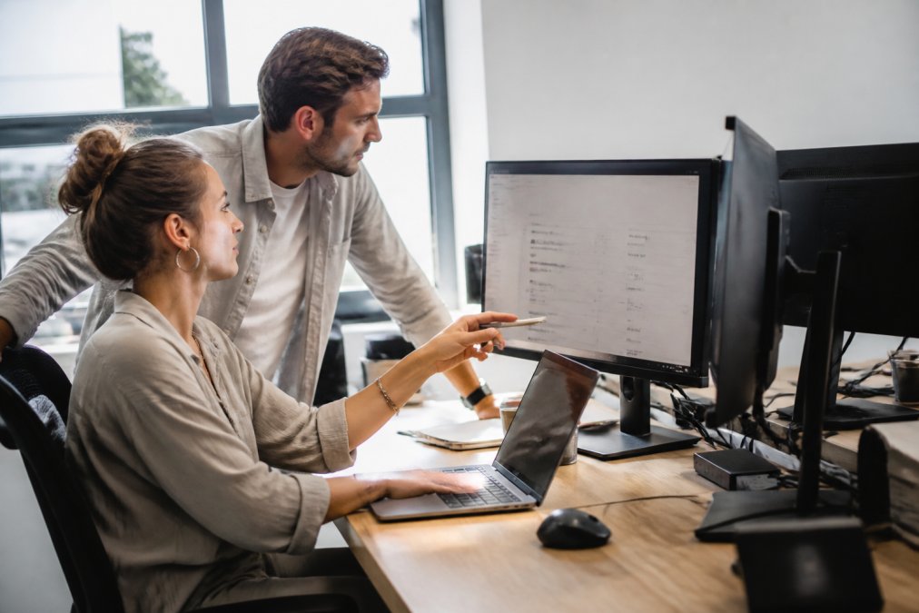 Professional working at desk with multiple monitors displaying business software dashboards and analytics interfaces in bright office environment