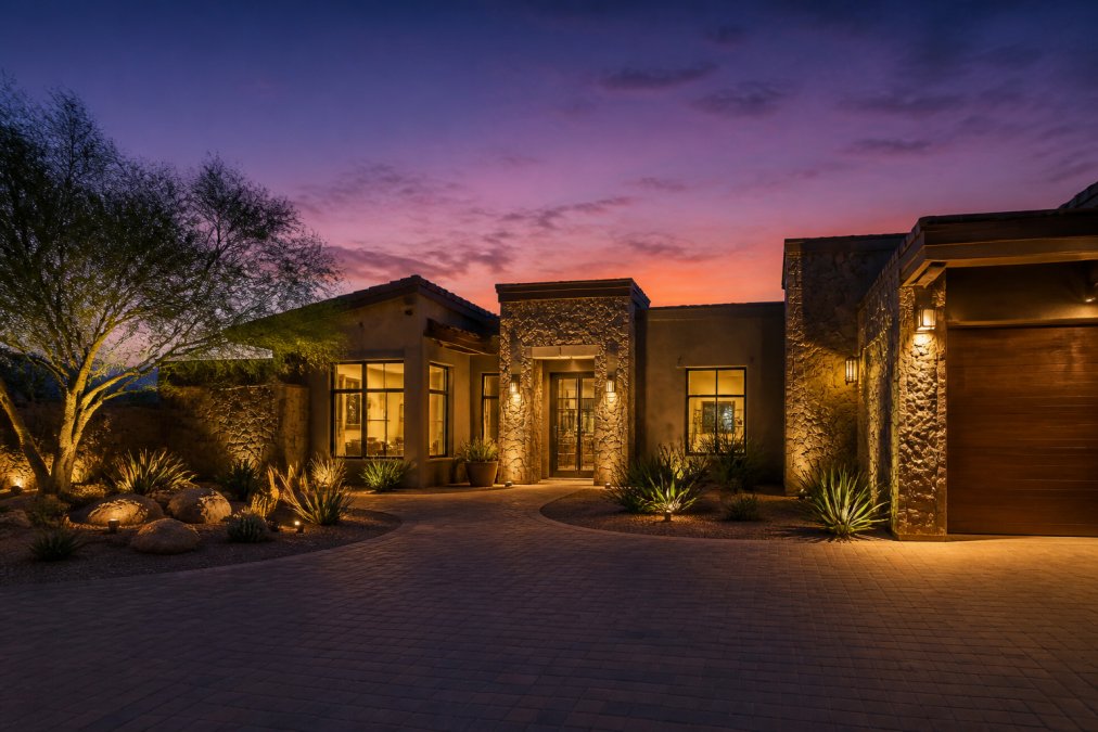 Luxury home exterior at dusk with warm architectural lighting illuminating the facade, glowing landscape features, and a sky transitioning from golden to deep purple, showcasing curb appeal and premium ambiance