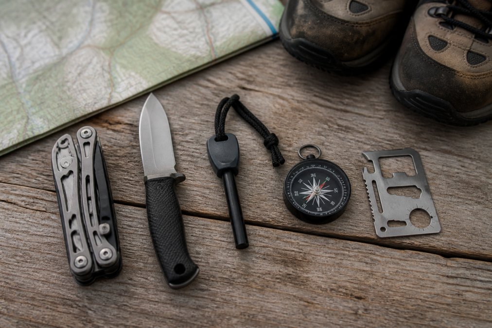 Overhead flat-lay of five survival tools arranged on weathered wood: a folding multi-tool, a fixed-blade knife, a ferro rod fire starter, a compact compass, and a credit-card-sized tool card, with a topographic map and hiking boots in the background