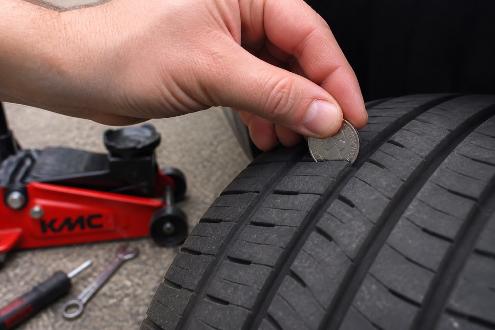A close-up of a hand pressing a 20p coin into the tread of a car tyre on a driveway in daylight, checking the depth against the coin's outer band