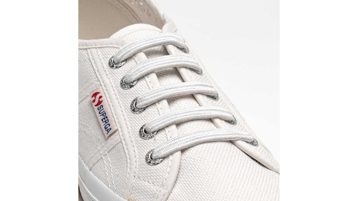 A parent's hands guiding a child's white sneaker while threading a white elastic no-tie lace through the eyelets, close-up view with soft natural light