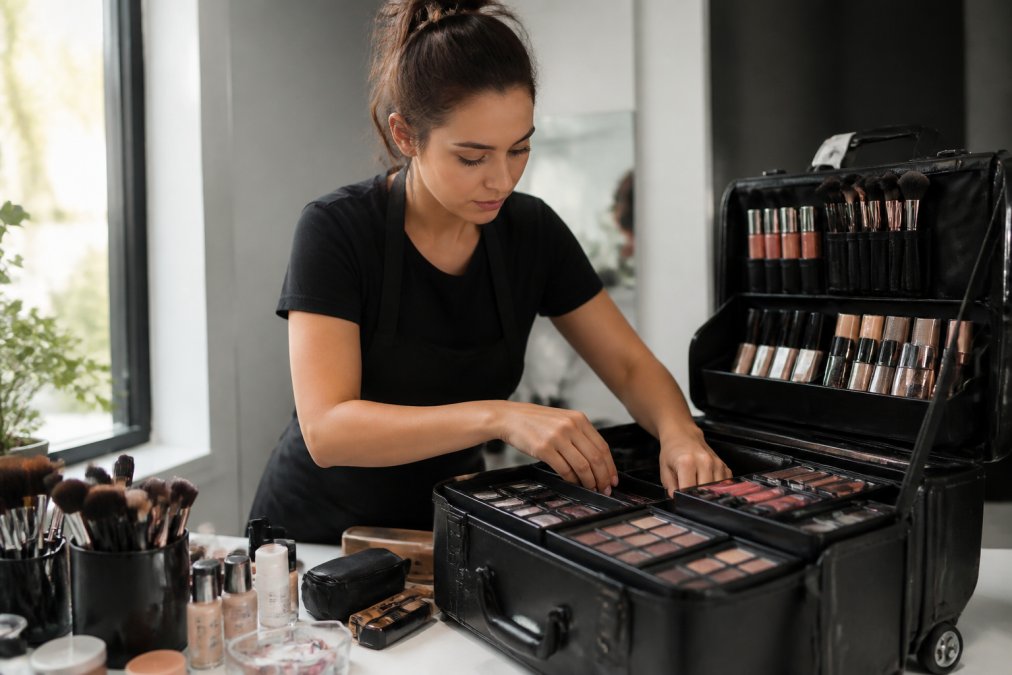 A professional makeup artist organizing a large rolling makeup case filled with palettes, brushes, and bottles on a clean studio counter, natural light from a window