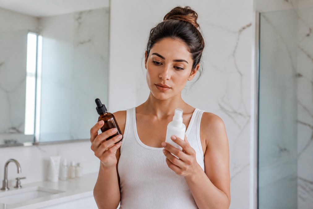 A woman in a bright bathroom holding two different skincare bottles side by side, comparing labels with a focused expression