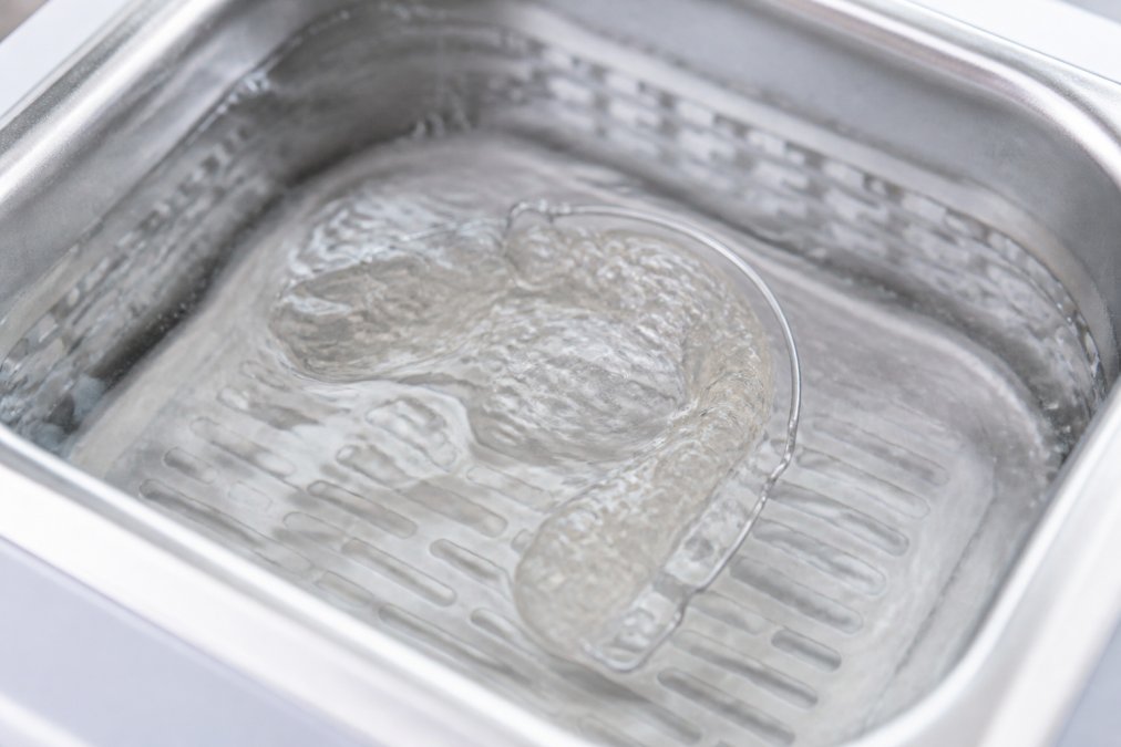 Nahaufnahme eines Retainers im Ultraschallbad mit sichtbaren Wasserwellen und feinen Bewegungen, die die Kavitation visualisieren, unter heller Beleuchtung