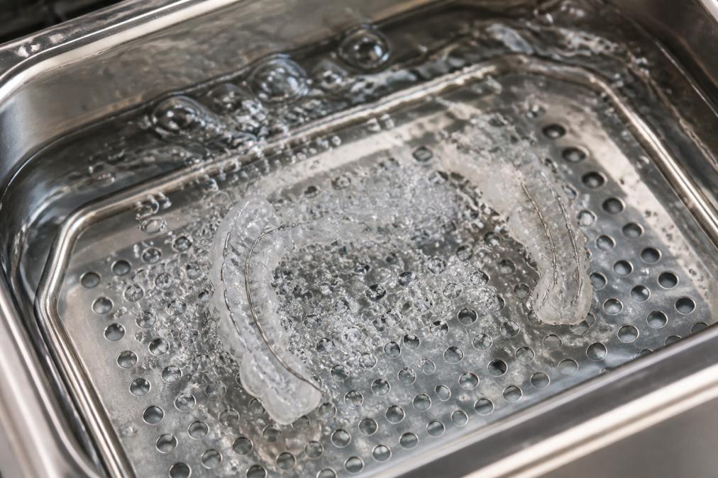 Nahaufnahme eines Retainers in einem Ultraschallreinigungsbad mit sichtbaren Blasen und Wasserbewegung, die die Reinigungsaktion in Echtzeit zeigt