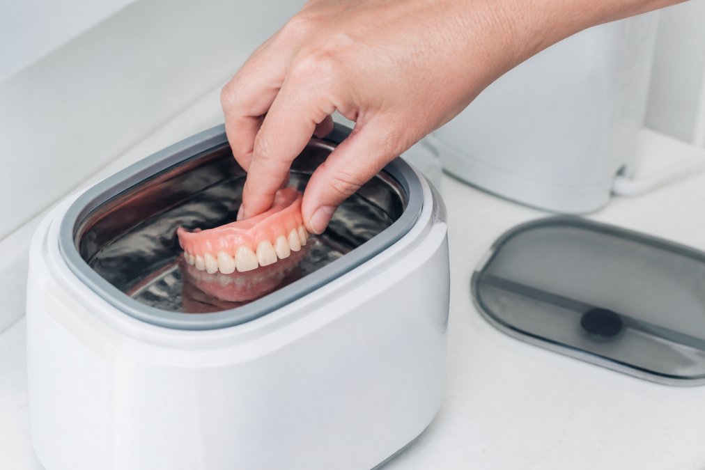 Nahaufnahme einer Hand, die eine Zahnprothese in ein Ultraschallreinigungsbad mit klarem Wasser einlegt, modernes Gerät mit Bedienfeld im Hintergrund
