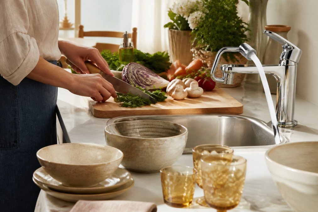 Close-up of hands preparing fresh vegetables on a wooden cutting board with artisan kitchen tools, ceramic serving bowls, and specialty glassware arranged on a marble countertop with warm natural light