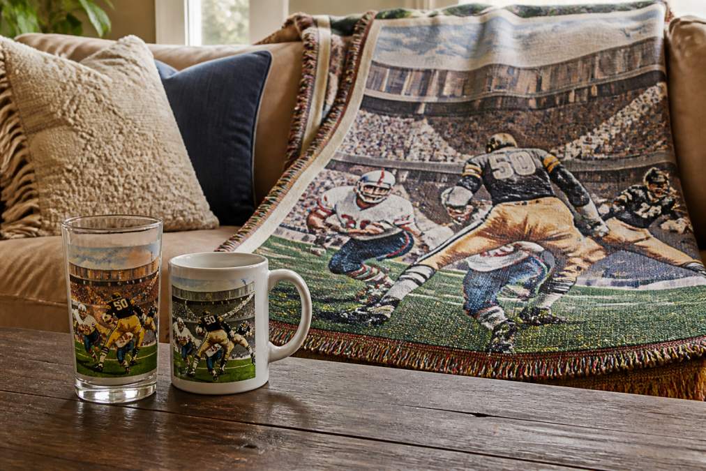 A handcrafted vintage sports blanket draped over a couch next to a mixing glass and ceramic mug with 1960s stadium artwork, showing personalized memorabilia gifts displayed in a cozy living space with warm natural lighting